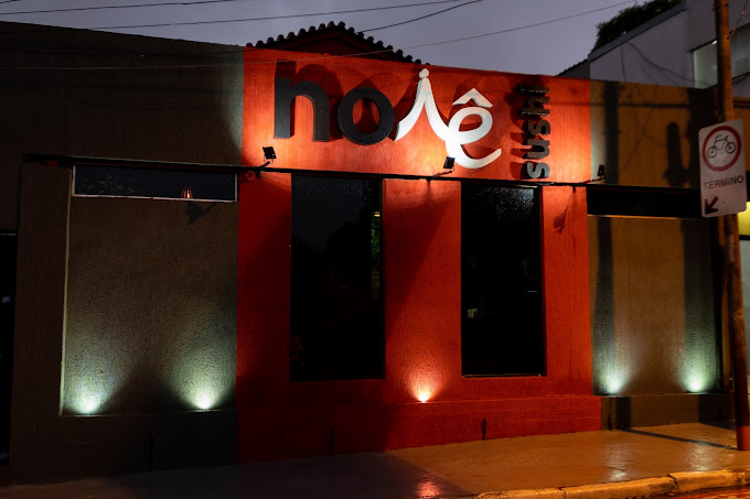 Fachada do Noiê Sushi iluminada à noite, com letreiro e parede vermelha